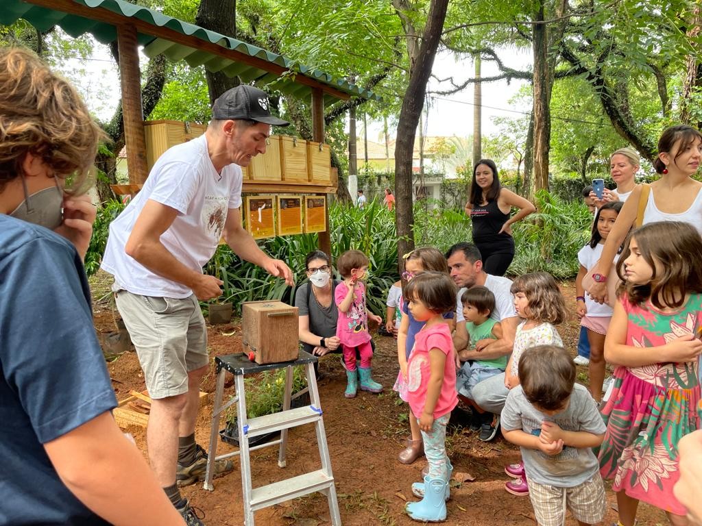 Visita de crianças e adultos ao meliponário da ANMA na Pracinha da Inhambu: um apicultor apresenta uma caixinha de abelhas sem ferrão apoiada num escadinho de alumínio, em frente ao abrigo de madeira das colmeias