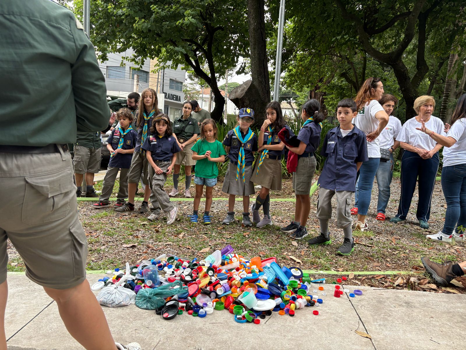 Crianças e escoteiros observando uma pilha de tampinhas plásticas separadas no chão da praça